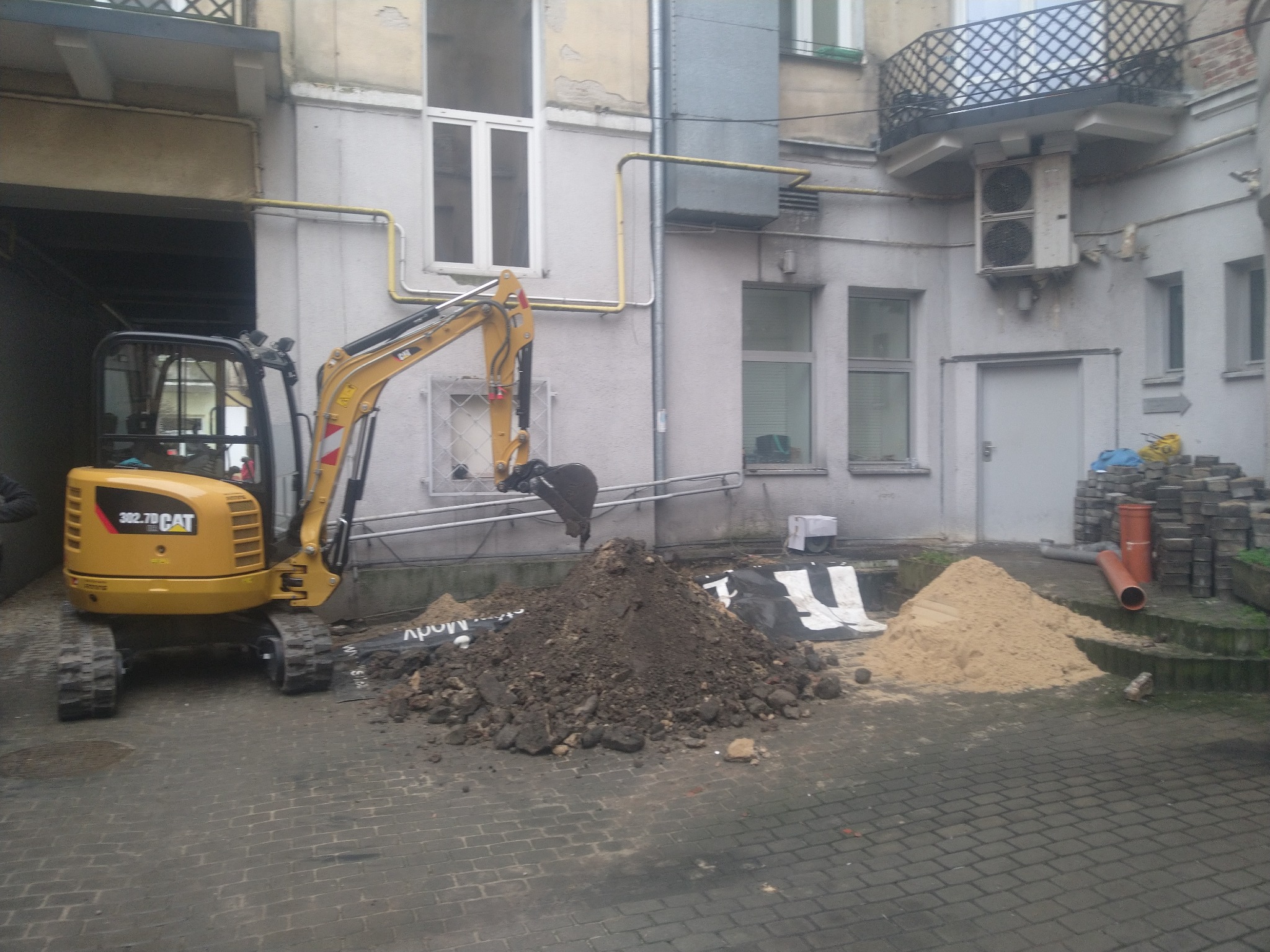 Żółta minikoparka CAT 302.7D nabiera ziemię na tle budynku z oknami i balkonem, obok leżą pryzmy ziemi i piasku.