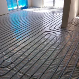 Szare rury instalacji podłogowej ułożone na folii z nadrukowaną siatką, widok z góry pod kątem, częściowo widoczne okno i ściana.