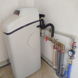 SANTERM - Zainstalowany zmiękczacz wody z widocznym panelem sterowania, podłączony do rozbudowanej sieci rur z mosiężnymi zaworami i niebieskim wodomierzem, na tle jasnej ściany.