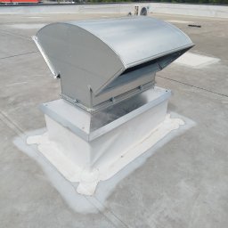 SANTERM - Metalowa, łukowata czerpnia dachowa zamontowana na białej podstawie, szczelnie uszczelniona na szarym płaskim dachu. W tle miejski krajobraz z zielenią.