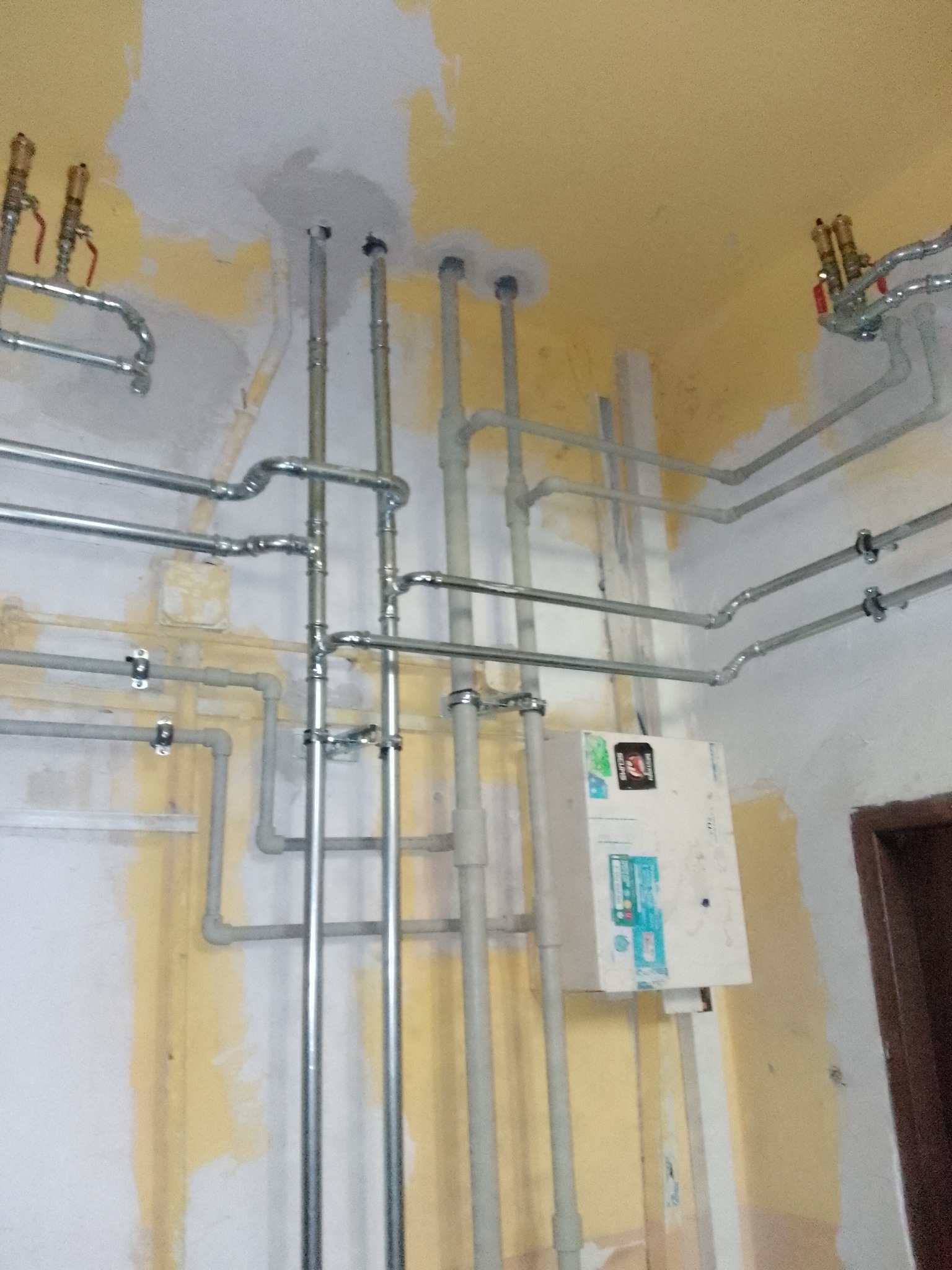 Szereg rur instalacyjnych, zarówno pionowych, jak i poziomych, z zaworami, zamontowanych na niedokończonej ścianie z resztkami farby. Widoczna skrzynka z naklejkami.