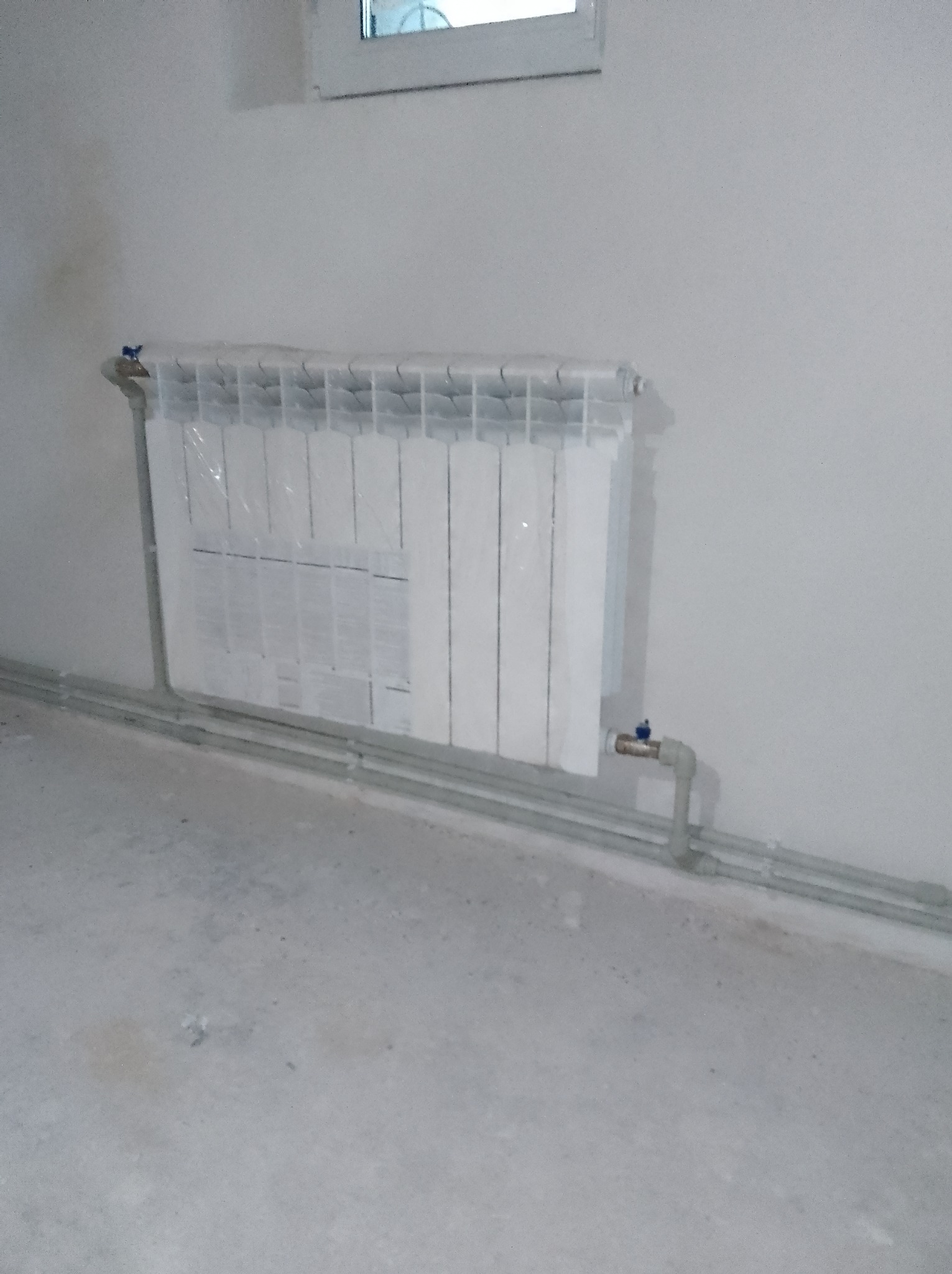 Biały, aluminiowy grzejnik panelowy, jeszcze w ochronnej folii, zamontowany na białej ścianie. Widoczne szare rury doprowadzające wodę oraz zawór termostatyczny.