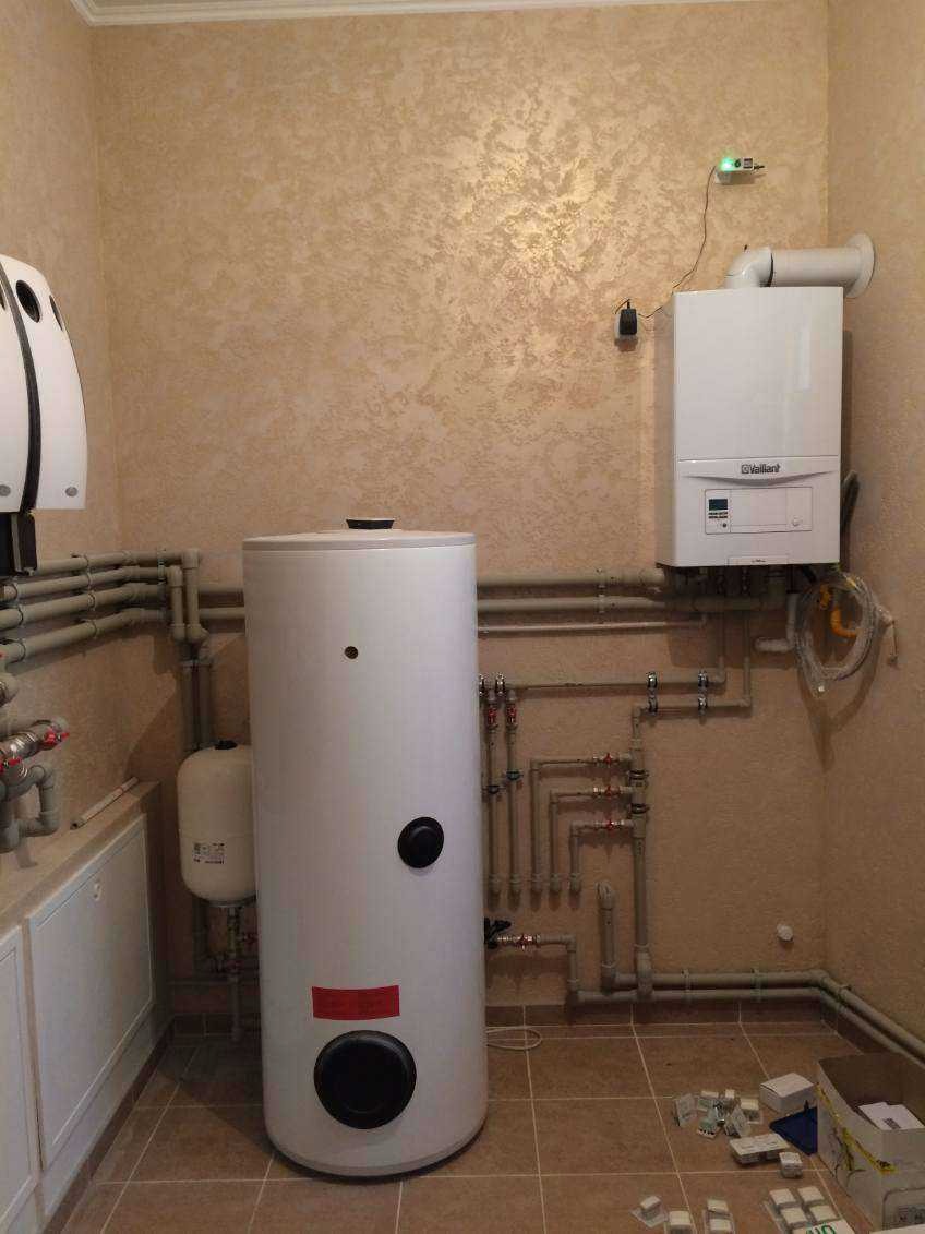Biały zasobnik ciepłej wody użytkowej z kotłem gazowym Vaillant i rurami w pomieszczeniu o beżowych ścianach i brązowej podłodze. Widoczne liczne przyłącza.