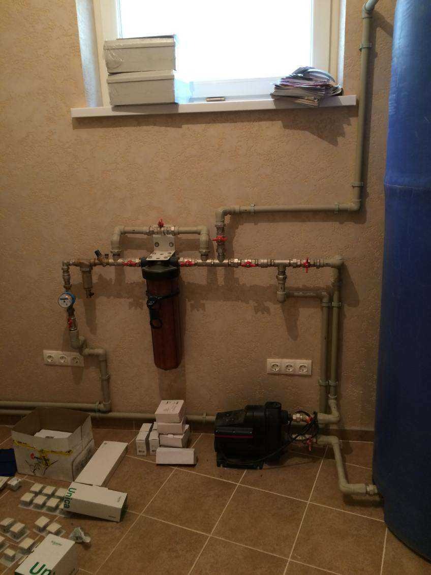 Złożona instalacja wodna z filtrem, pompą i zbiornikiem hydroforowym w pomieszczeniu z beżowymi ścianami i brązową podłogą. Widoczne liczniki, zawory i elementy montażowe.