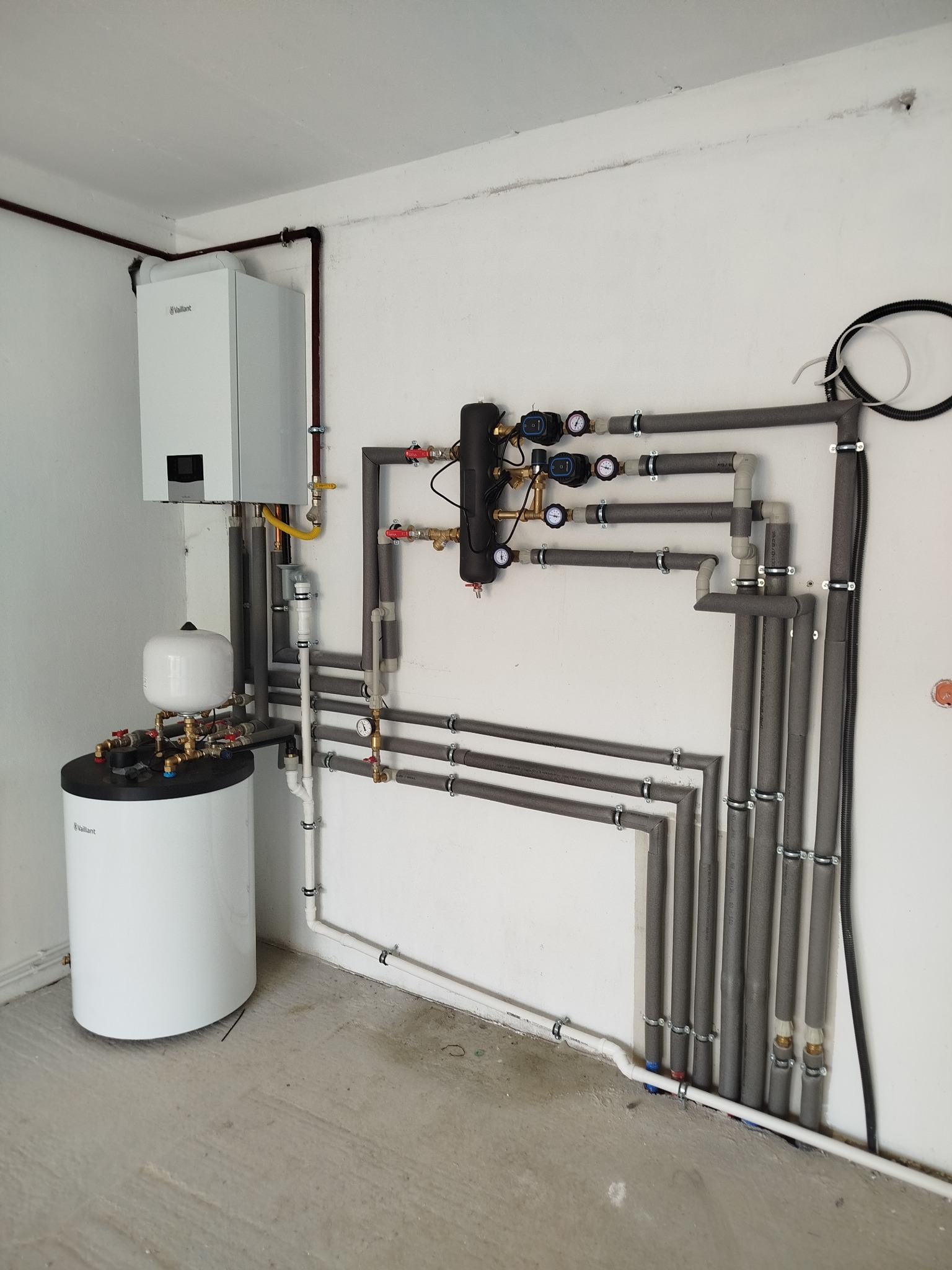 Biały piec i zasobnik Vaillant z szarymi rurami w izolacji, manometry i zawory na białej ścianie. Instalacja grzewcza w kotłowni, widok z dołu.