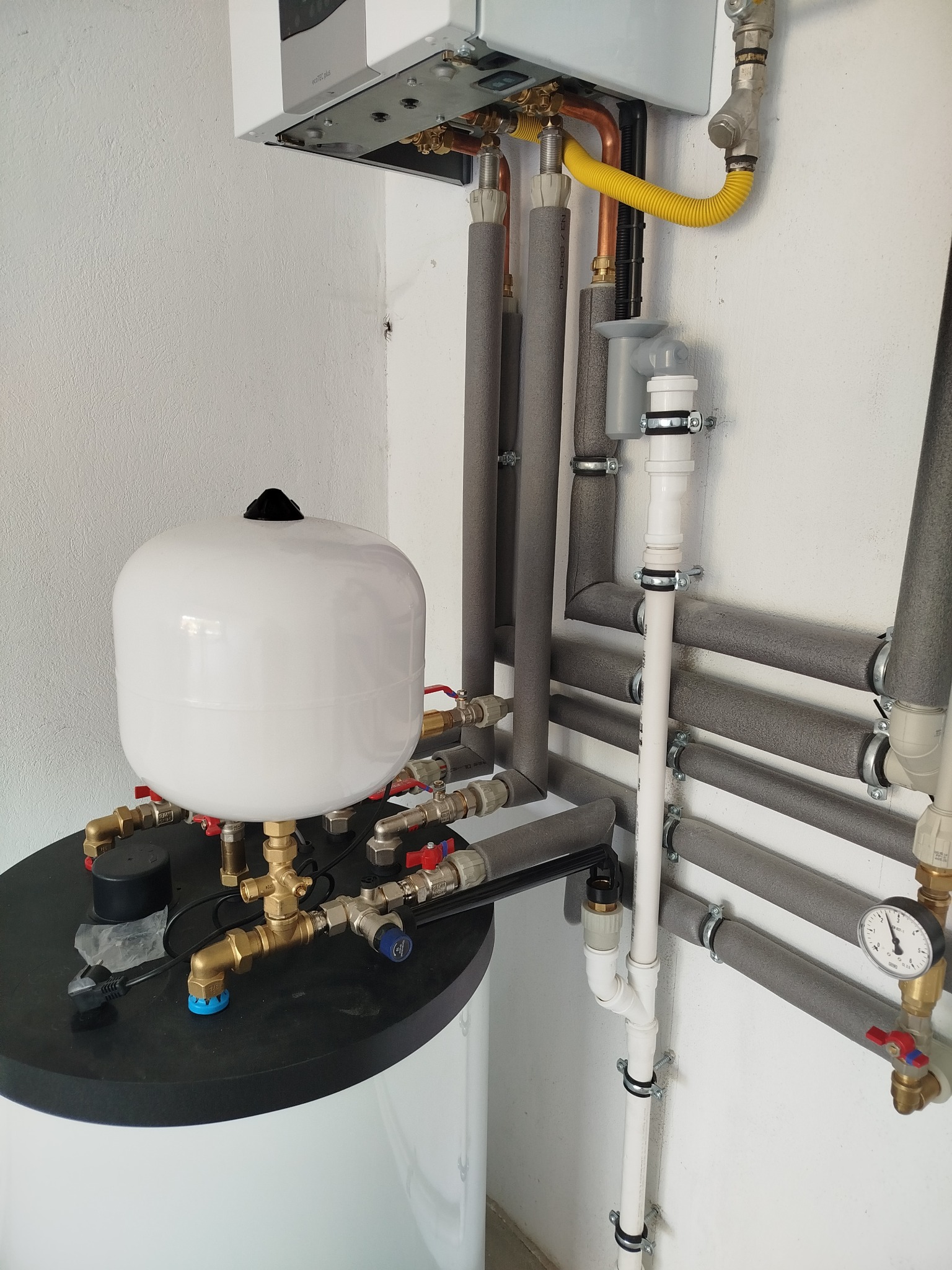 Złożona instalacja grzewcza z białym zbiornikiem wyrównawczym, rurami w izolacji termicznej i kotłem wiszącym na ścianie. Widoczne liczne zawory i manometr.