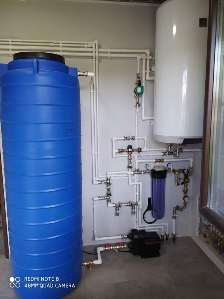 Kompleksowa instalacja: niebieski zbiornik akumulacyjny, biały bojler, filtr wody i pompa obiegowa połączone rurami. Widok na szare ściany i podłogę w pomieszczeniu.