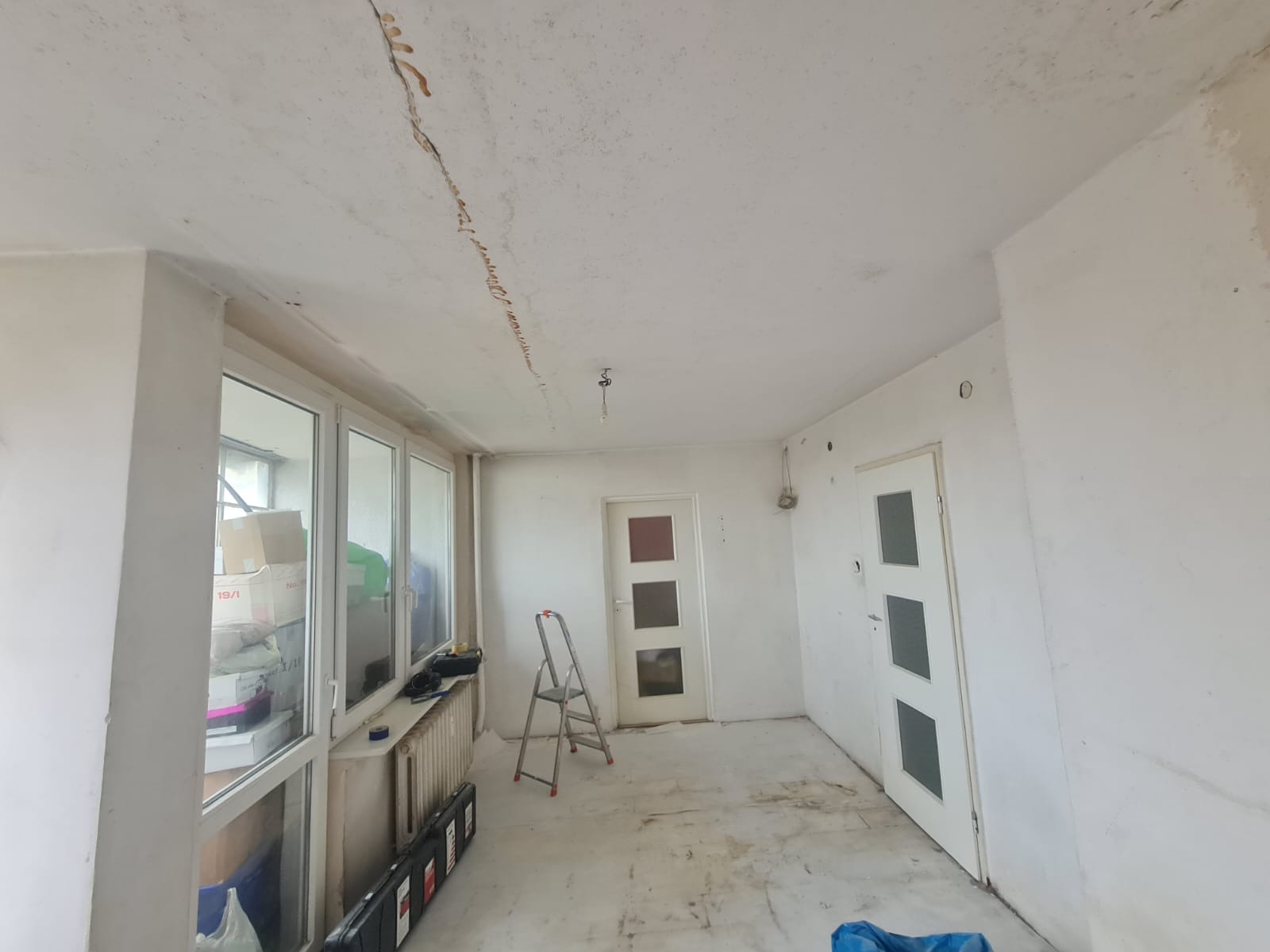 Pokój w trakcie przygotowania do remontu, widoczne pęknięcie na suficie, drabina, okna z kartonami, podłoga zabezpieczona folią.