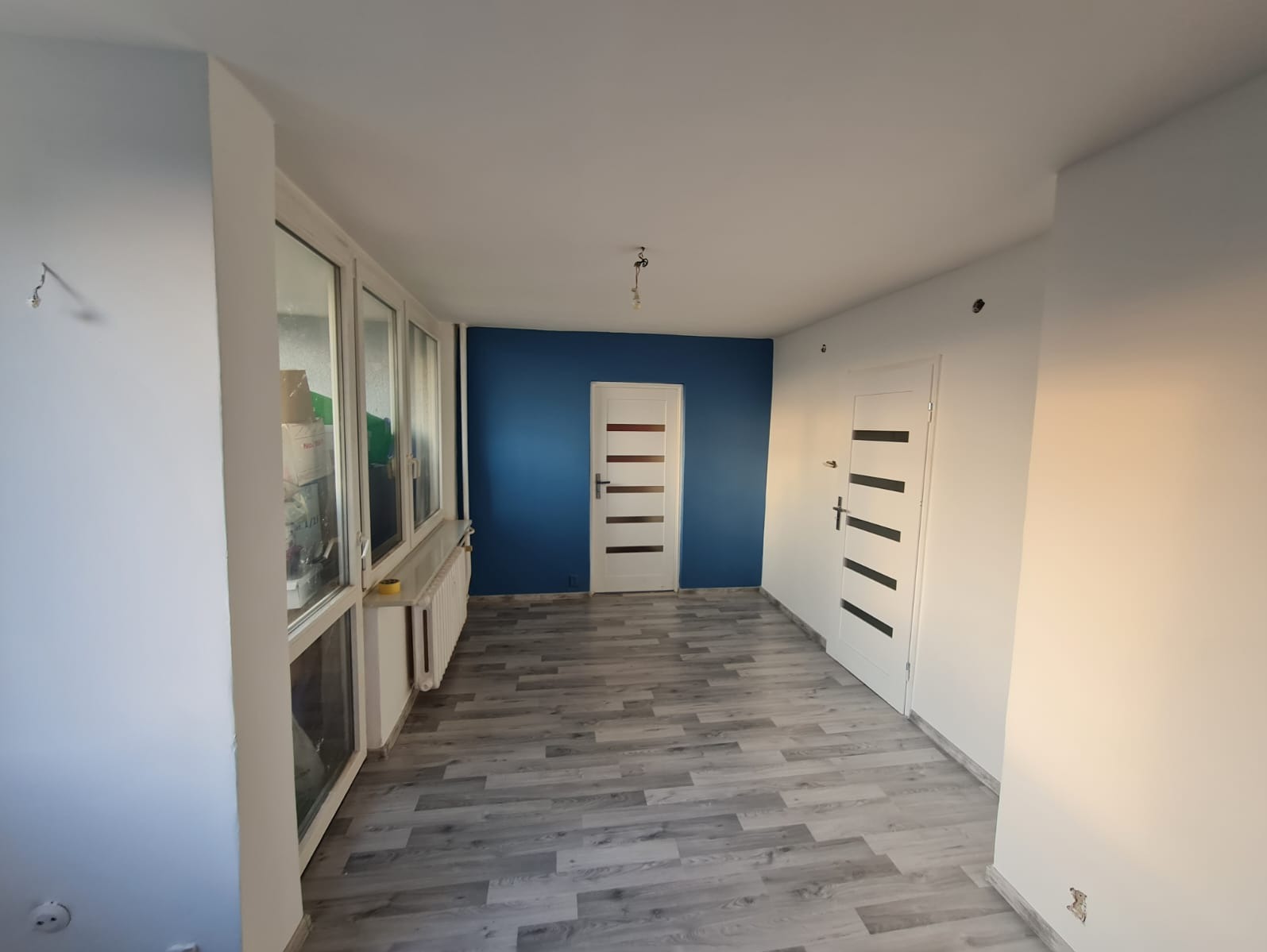 Pomieszczenie po remoncie: błękitna ściana, białe ściany, szara podłoga imitująca drewno, białe drzwi z poziomymi, czarnymi paskami, białe okna z widocznymi przedmiotami wewnątrz.