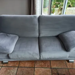 Dwuosobowa, szara sofa z weluru na niskich, chromowanych nóżkach, stojąca na brązowej, kwadratowej podłodze z płytek w jasnym wnętrzu.