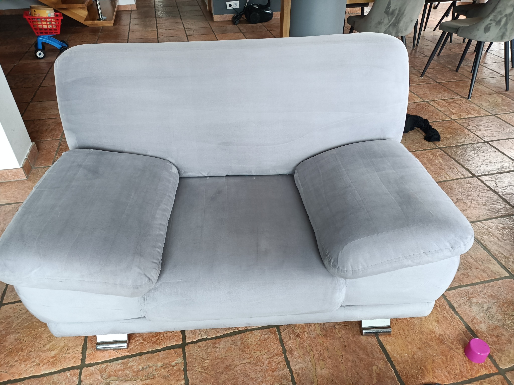 Szary, pluszowy fotel z podłokietnikami na kafelkowej podłodze. W tle zabawkowy wózek i krzesła. Widoczne ślady użytkowania na materiale.