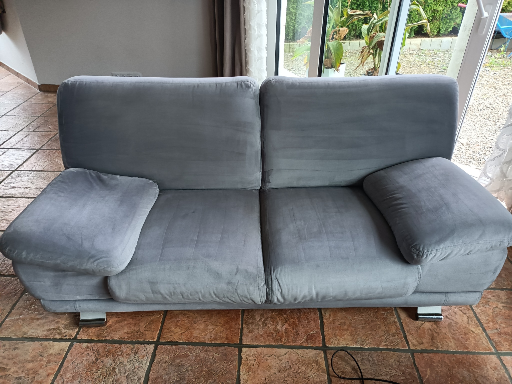 Dwuosobowa, szara sofa z weluru na niskich, chromowanych nóżkach, stojąca na brązowej, kwadratowej podłodze z płytek w jasnym wnętrzu.