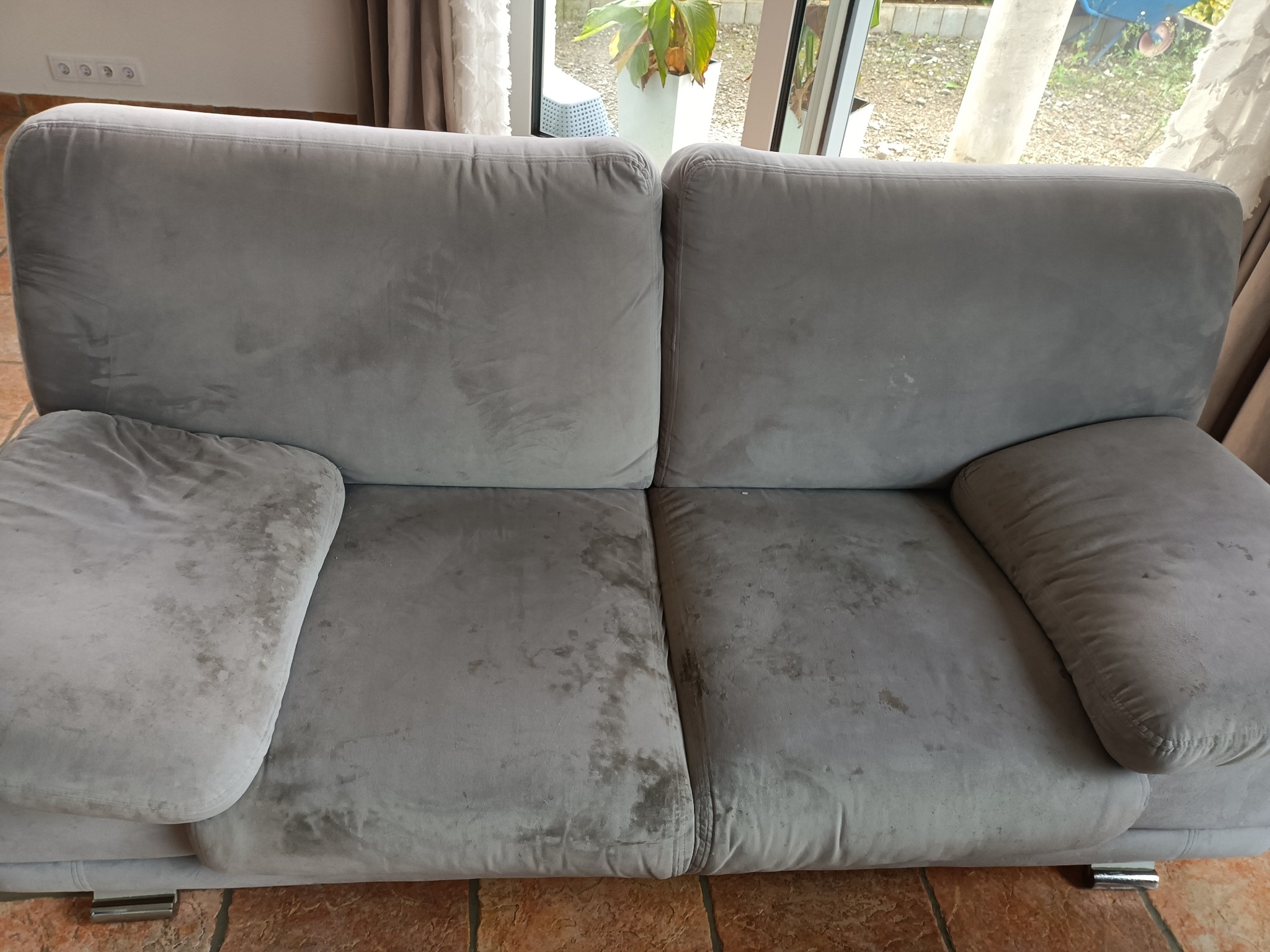 Brudna, szara sofa z plamami na siedzisku i podłokietnikach, widoczna na tle okna z roślinami. Wymaga czyszczenia tapicerki.