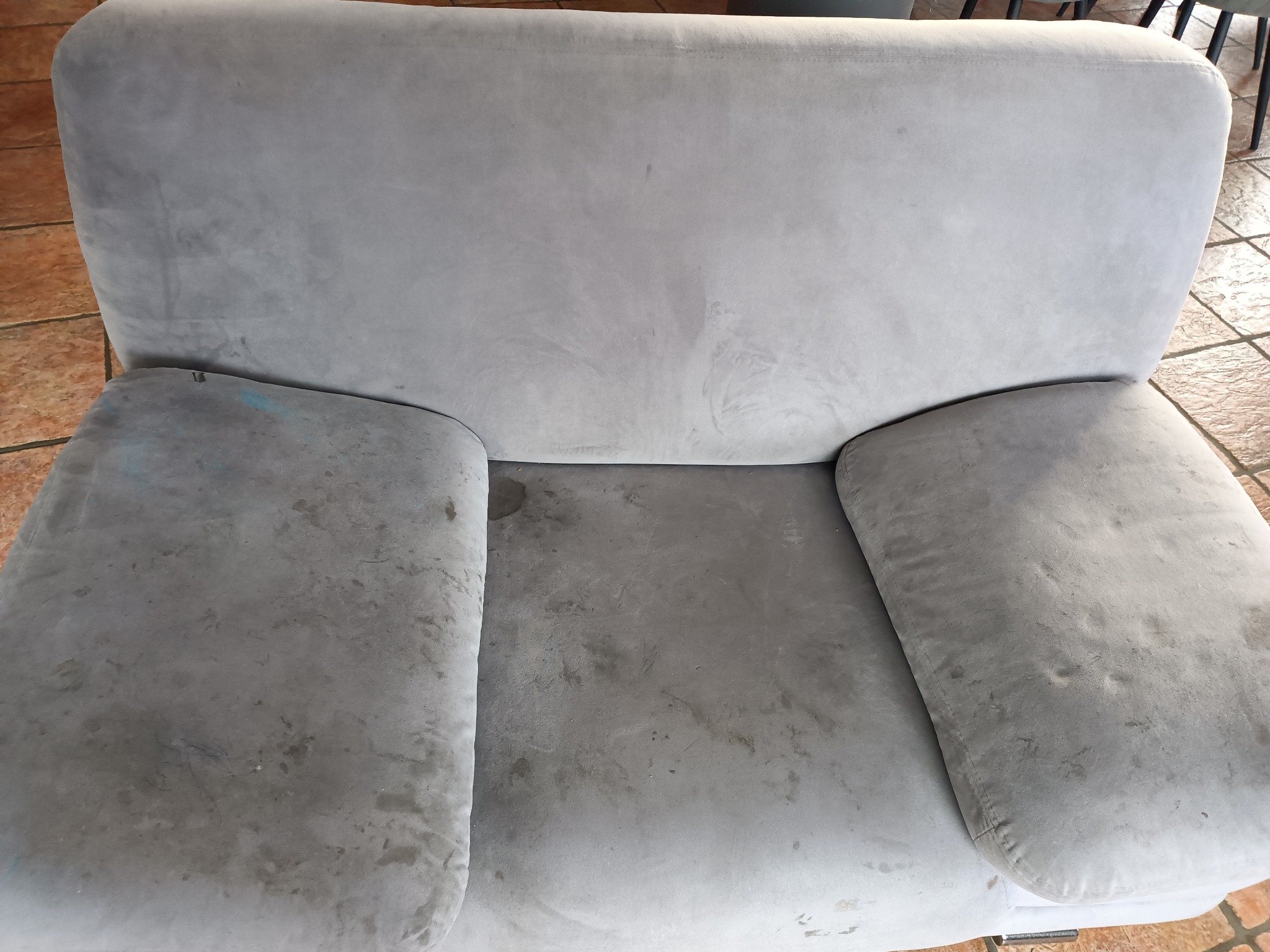 Szary, pluszowy fotel z widocznymi zabrudzeniami na siedzisku i podłokietnikach, czekający na profesjonalne czyszczenie tapicerki.