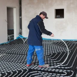 Instalator w niebieskiej kurtce i spodniach układa białe rury ogrzewania podłogowego na czarnych mocowaniach w surowym pomieszczeniu z widoczną instalacją elektryczną.