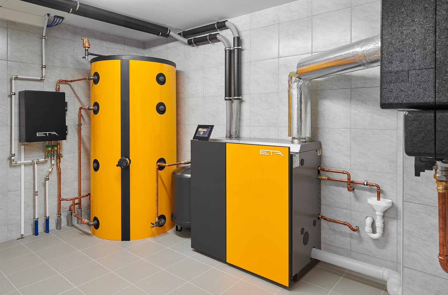 Nowoczesna kotłownia z żółto-szarym piecem, zbiornikiem buforowym i instalacją hydrauliczną z miedzianych rur, w pomieszczeniu wykończonym płytkami ceramicznymi.