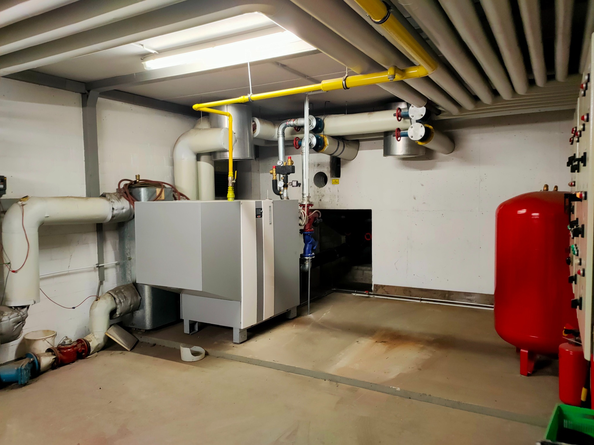 Węzeł cieplny w piwnicy z szarym piecem, czerwonym zbiornikiem wyrównawczym, żółtymi rurami gazowymi pod sufitem i licznymi rurami grzewczymi