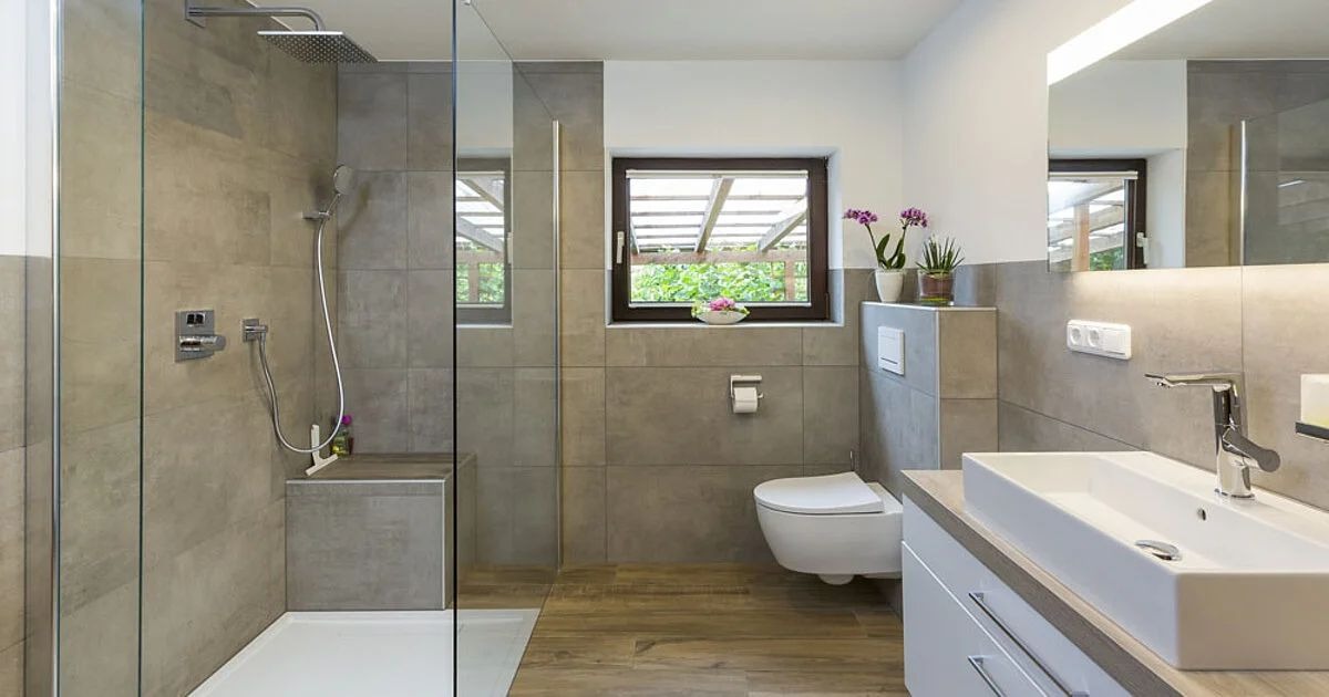 Nowoczesna łazienka z przeszkloną kabiną prysznicową typu walk-in, wiszącą toaletą i umywalką nablatową z chromowaną armaturą, wykończona szarą płytką i drewnopodobną podłogą.