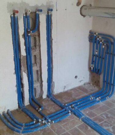 Niebieskie rury instalacyjne, poprowadzone w bruzdach w ścianie i na podłodze, z licznymi złączkami, w surowym stanie wykończenia wnętrza.