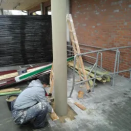 Pracownik w szarej kurtce układa kostkę brukową wokół betonowego słupa na zewnątrz budynku, widoczna drabina oparta o słup i ceglana ściana w tle.