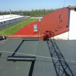 Widok z góry na dach pokryty czarną papą, z rolkami papy i cieniem masztu antenowego, w tle czerwony dach budynku przemysłowego i zieleń drzew.