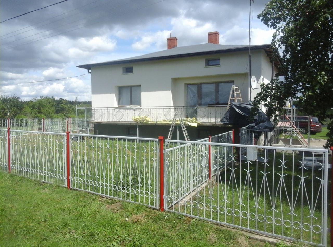 Dwupiętrowy dom z nowym dachem i biało-czerwonym ogrodzeniem, widoczne rusztowania i materiały zabezpieczone folią, sugerujące trwające prace wykończeniowe.