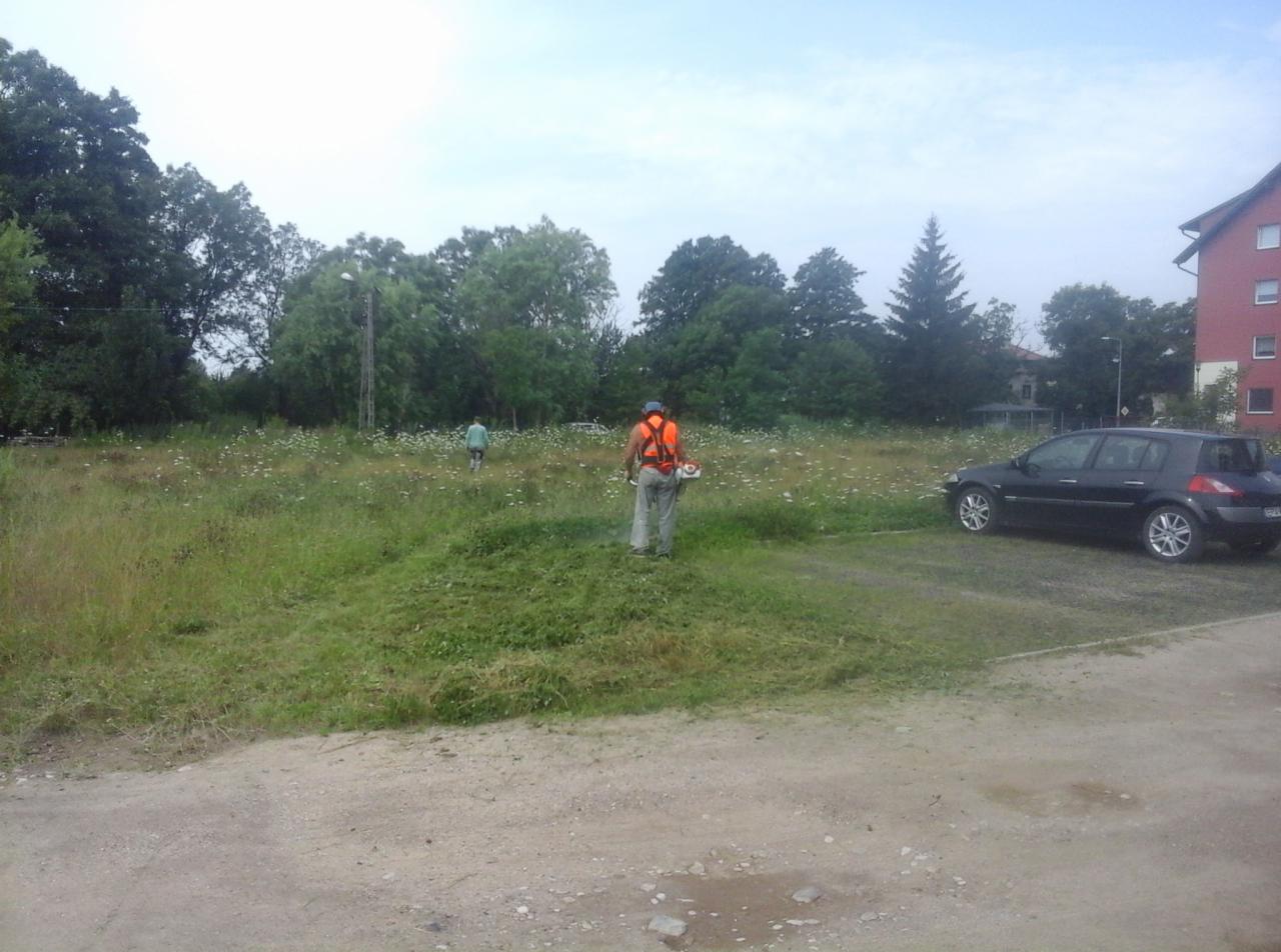 Pracownik w pomarańczowej kamizelce tnie trawę kosiarką spalinową na zarośniętej działce, w tle zaparkowany samochód i budynek mieszkalny.