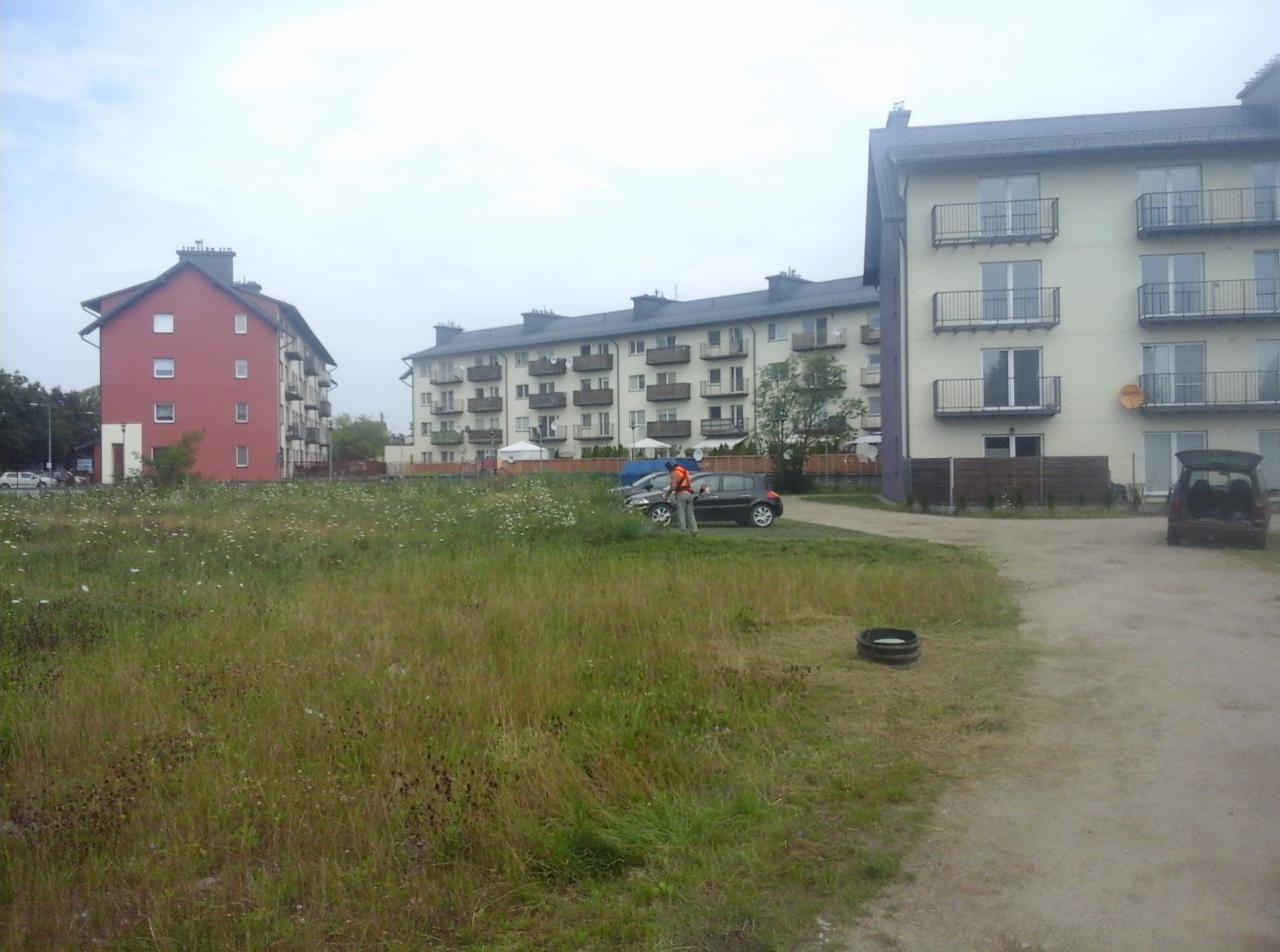 Mężczyzna w kamizelce odblaskowej kosi trawę kosiarką spalinową na zarośniętym terenie w Głownie, na tle budynków mieszkalnych z balkonami.