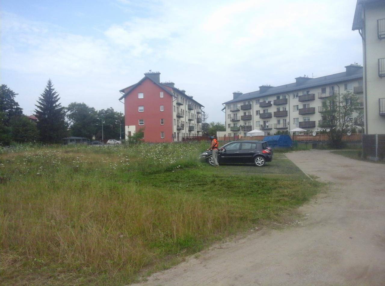 Mężczyzna w odblaskowej kamizelce kosi trawę przy samochodzie na tle bloków mieszkalnych i zarośniętego pola.