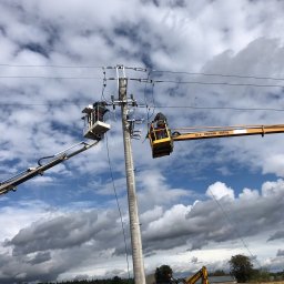 MODERNITY Michał Lech - Prace na słupie energetycznym: dwóch elektryków w podnośnikach koszowych, jeden w białym, drugi w żółtym, montują lub naprawiają instalację elektryczną na tle pochmurnego nieba.