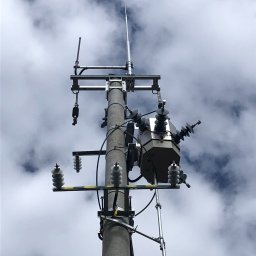 MODERNITY Michał Lech - Słup energetyczny z transformatorem i odgromnikiem na tle pochmurnego nieba, widok z dołu.