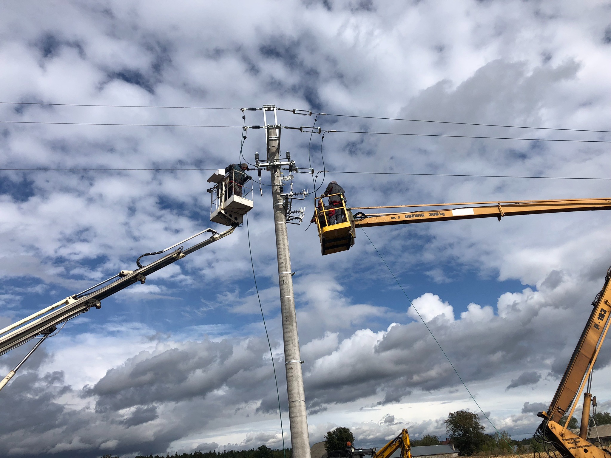 Prace na słupie energetycznym: dwóch elektryków w podnośnikach koszowych, jeden w białym, drugi w żółtym, montują lub naprawiają instalację elektryczną na tle pochmurnego nieba.