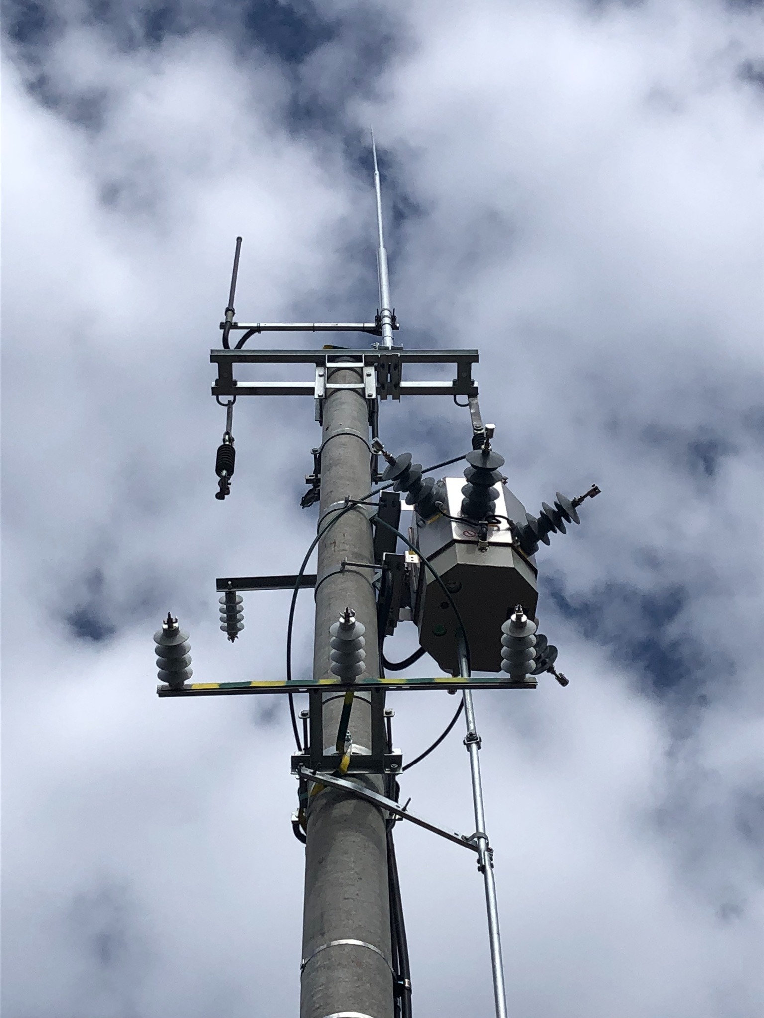 Słup energetyczny z transformatorem i odgromnikiem na tle pochmurnego nieba, widok z dołu.