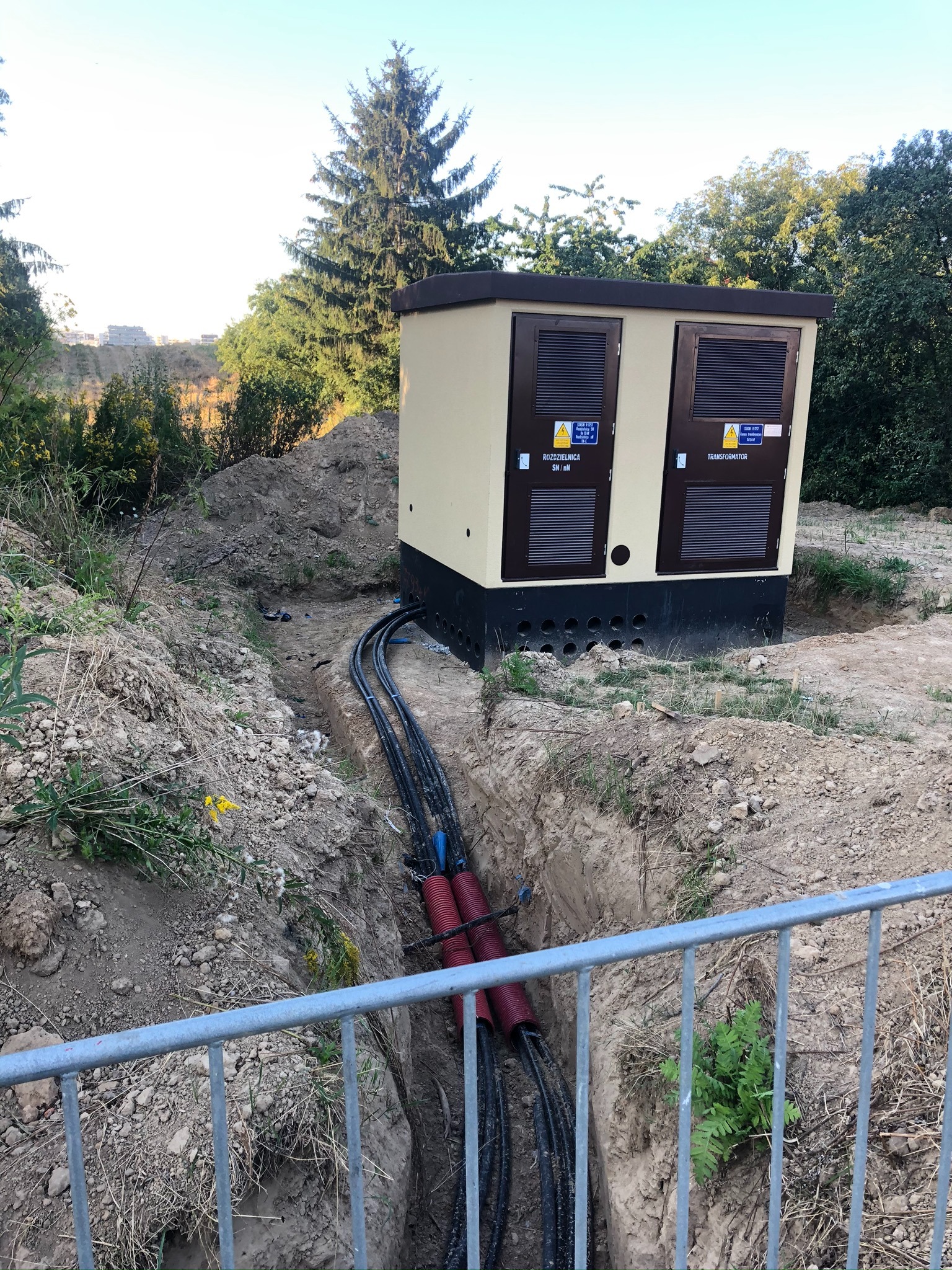 Zewnętrzna stacja transformatorowa z podłączonymi kablami w wykopie, zabezpieczona metalowym ogrodzeniem.