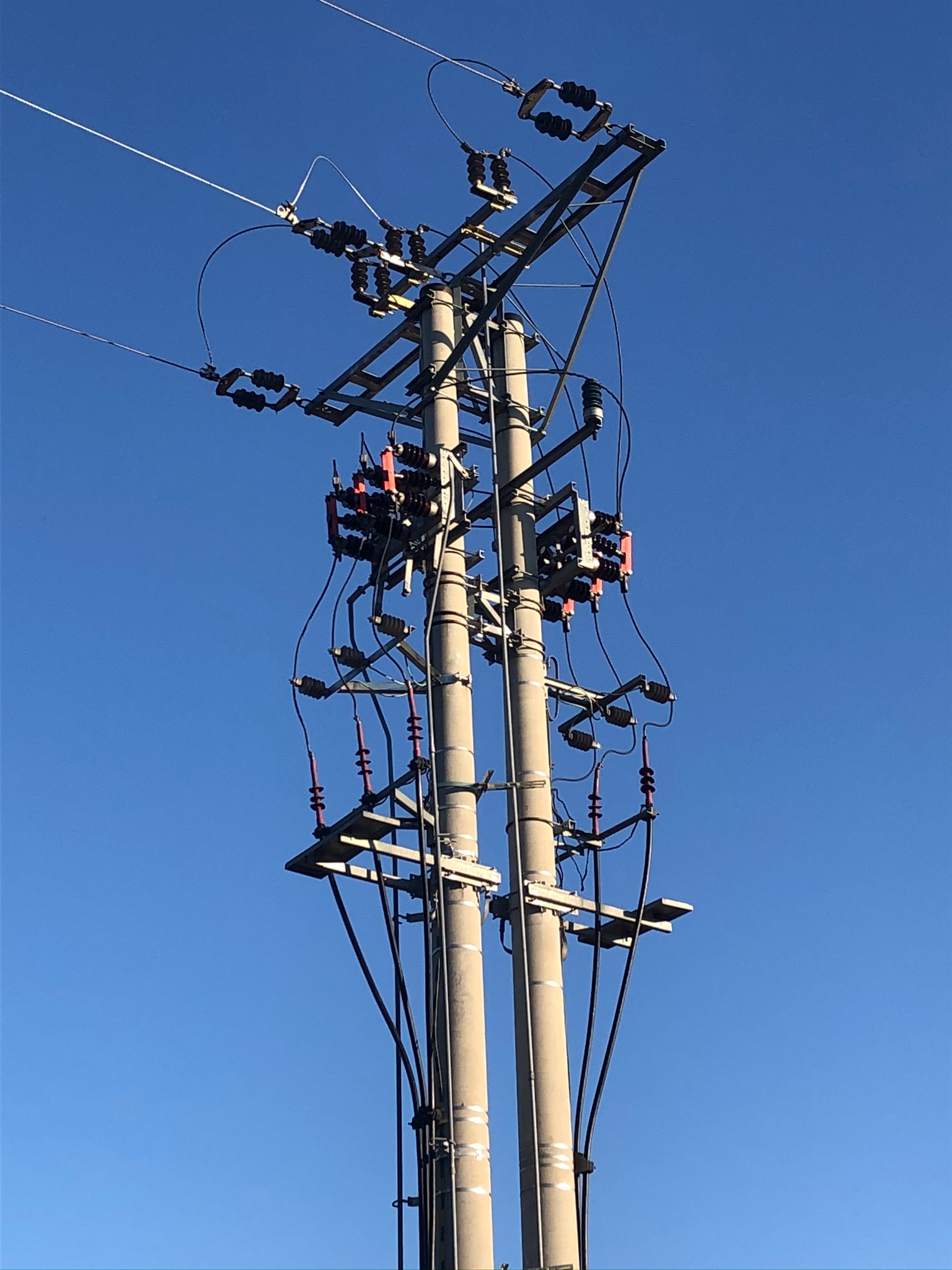 Słup wysokiego napięcia z przewodami i izolatorami na tle błękitnego nieba, widoczny z dołu.