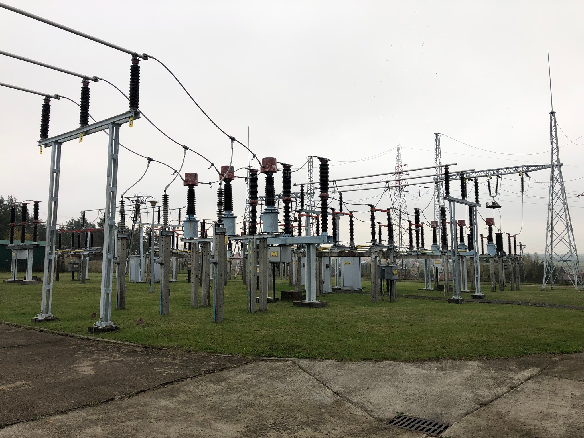 Stacja transformatorowa z rozległym układem przewodów i izolatorów, widoczna z poziomu gruntu, z metalową kratką ściekową w prawym dolnym rogu.