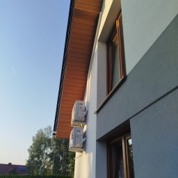 MK KLIMWENT SPÓŁKA Z OGRANICZONĄ ODPOWIEDZIALNOŚCIĄ - Dwa klimatyzatory zamontowane na elewacji domu z drewnianym podbitkiem, widok z dołu, błękitne niebo w tle.