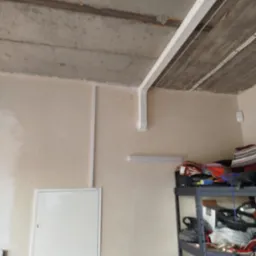 Białe rury wentylacyjne zamontowane pod betonowym sufitem w pomieszczeniu, widoczna szafka narzędziowa i skrzynka rewizyjna na ścianie.