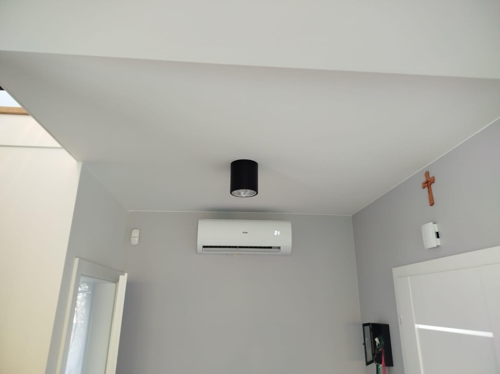 Biały klimatyzator Haier zamontowany na szarej ścianie w pokoju, widoczny czarny plafon na suficie i drewniany krzyż na ścianie obok drzwi.