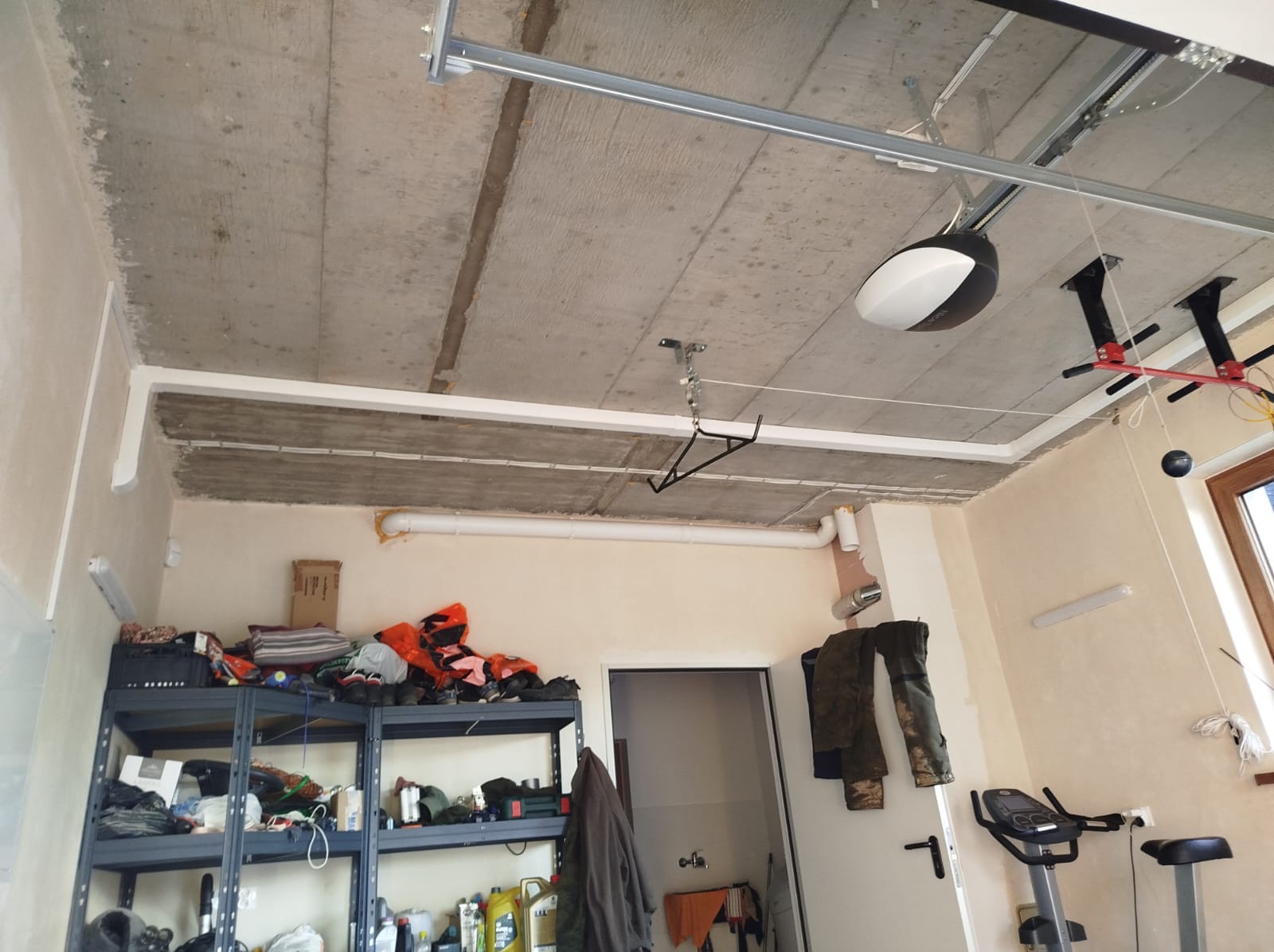 Montaż automatyki bramy garażowej: widoczny napęd pod sufitem, surowy betonowy strop, instalacja wentylacyjna z rur PCV, regał z narzędziami i odzieżą roboczą w garażu.