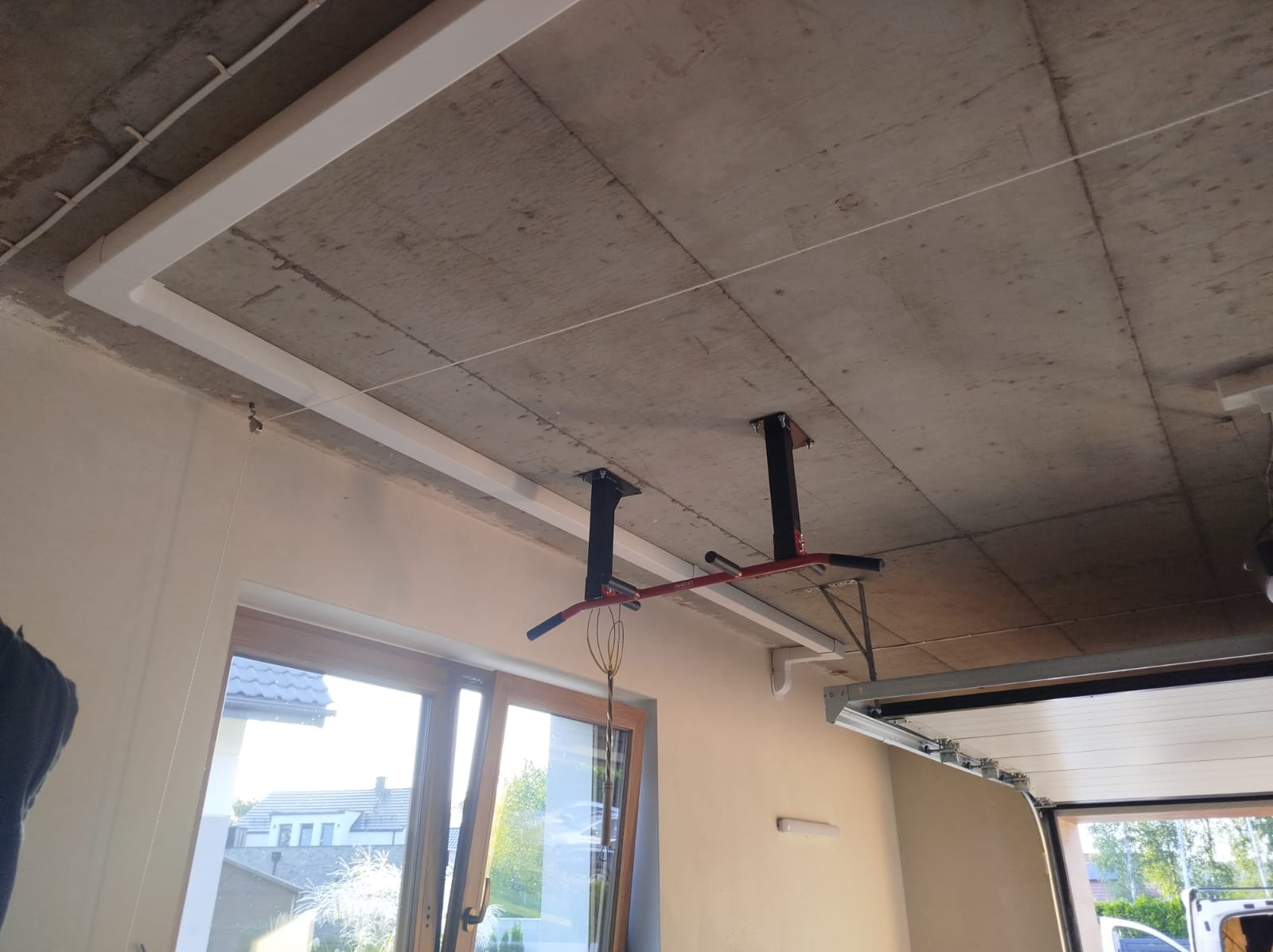 Surowy betonowy sufit garażu z zamontowanymi białymi listwami maskującymi instalacje, oraz konstrukcją do ćwiczeń podciągania, widok na częściowo otwarte drzwi garażowe i okno.