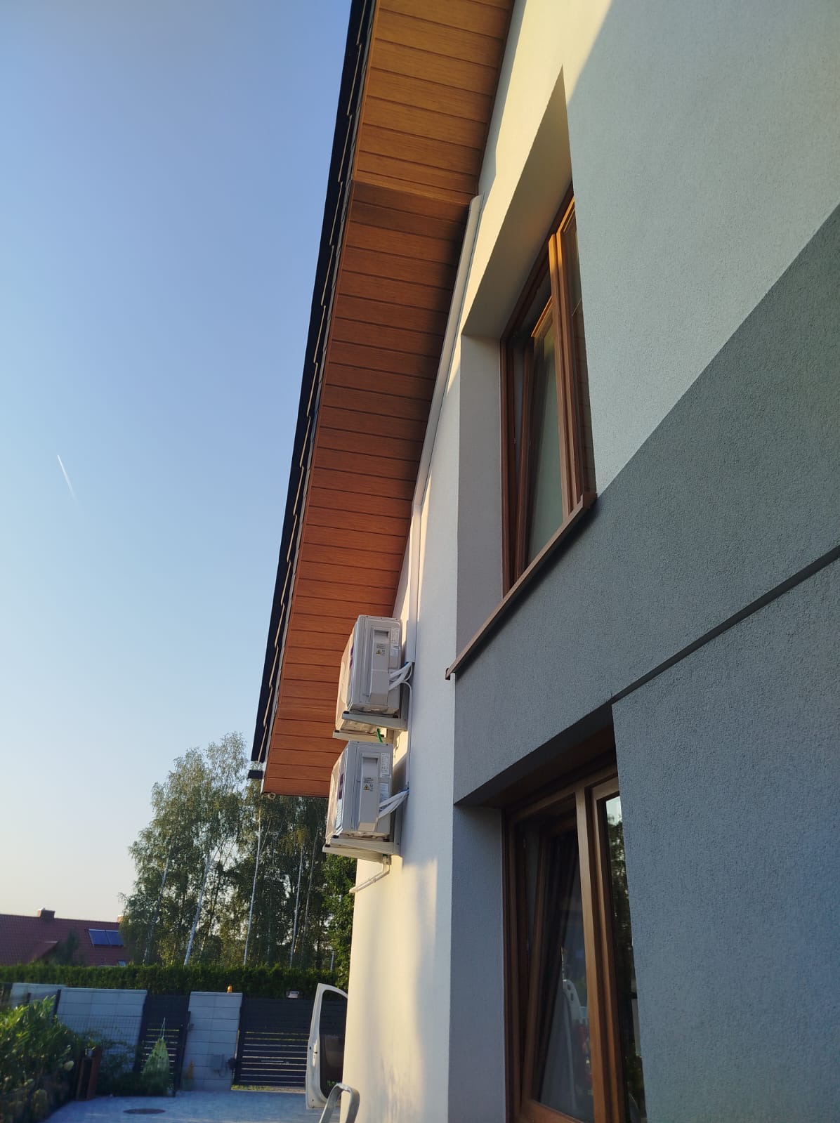 Dwa klimatyzatory zamontowane na elewacji domu z drewnianym podbitkiem, widok z dołu, błękitne niebo w tle.