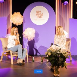 Jakub Dobkowski SlowLove Studio - J.Senses Sound & Vision - Dwie kobiety siedzą na scenie podczas konferencji, oświetlonej fioletowymi reflektorami, z dekoracjami kwiatowymi i butelkami wody mineralnej na podłodze.