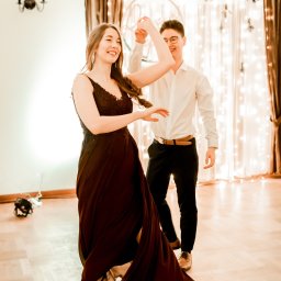 Jakub Dobkowski SlowLove Studio - J.Senses Sound & Vision - Uśmiechnięta para tańczy na parkiecie, kobieta w bordowej sukni z koronkową górą i baletkach, mężczyzna w białej koszuli i czarnych spodniach, w tle dekoracja ze światełek.