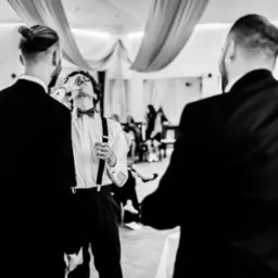 Mężczyzna w muszce i szelkach wznosi toast podczas uroczystości, widziany z perspektywy dwóch osób w garniturach, tło z dekoracjami.