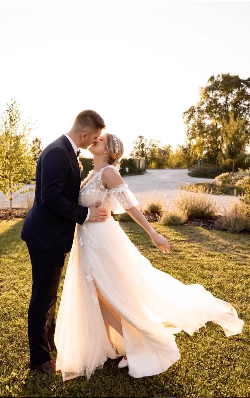 Pocałunek młodej pary w plenerze, suknia ślubna z rozcięciem i aplikacjami kwiatowymi, garnitur z muchą, złote światło zachodzącego słońca.