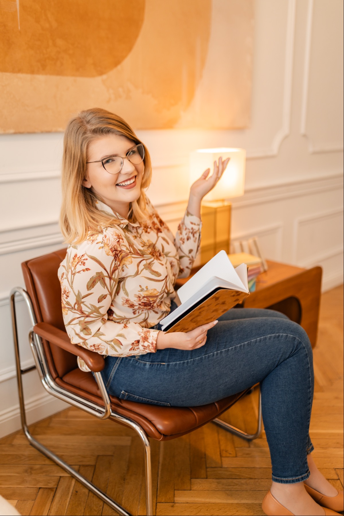 Uśmiechnięta blondynka w okularach siedzi w skórzanym fotelu, trzymając otwarty album ze złotą okładką i gestykulując ręką, w tle lampka i abstrakcyjny obraz.