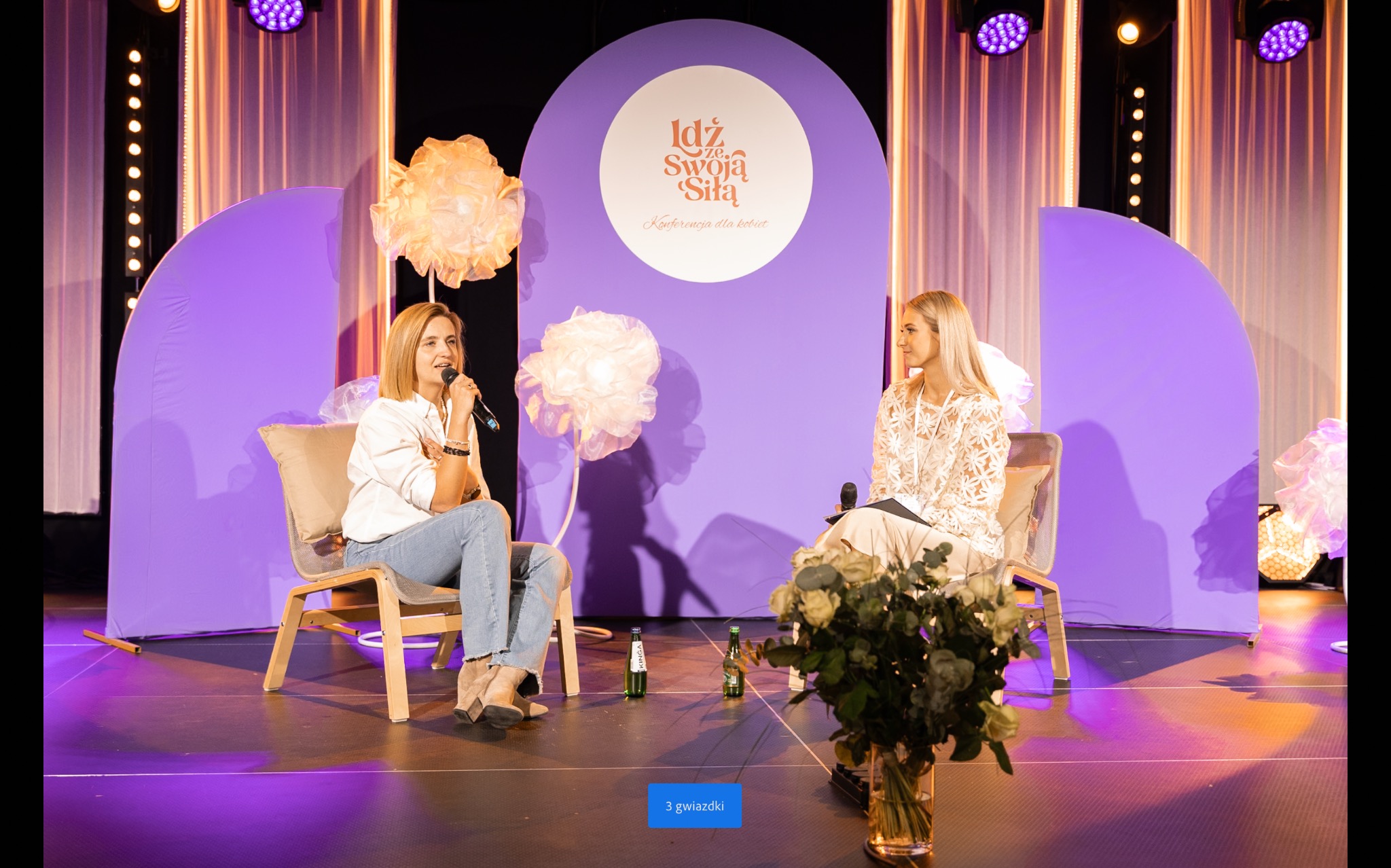 Dwie kobiety siedzą na scenie podczas konferencji, oświetlonej fioletowymi reflektorami, z dekoracjami kwiatowymi i butelkami wody mineralnej na podłodze.