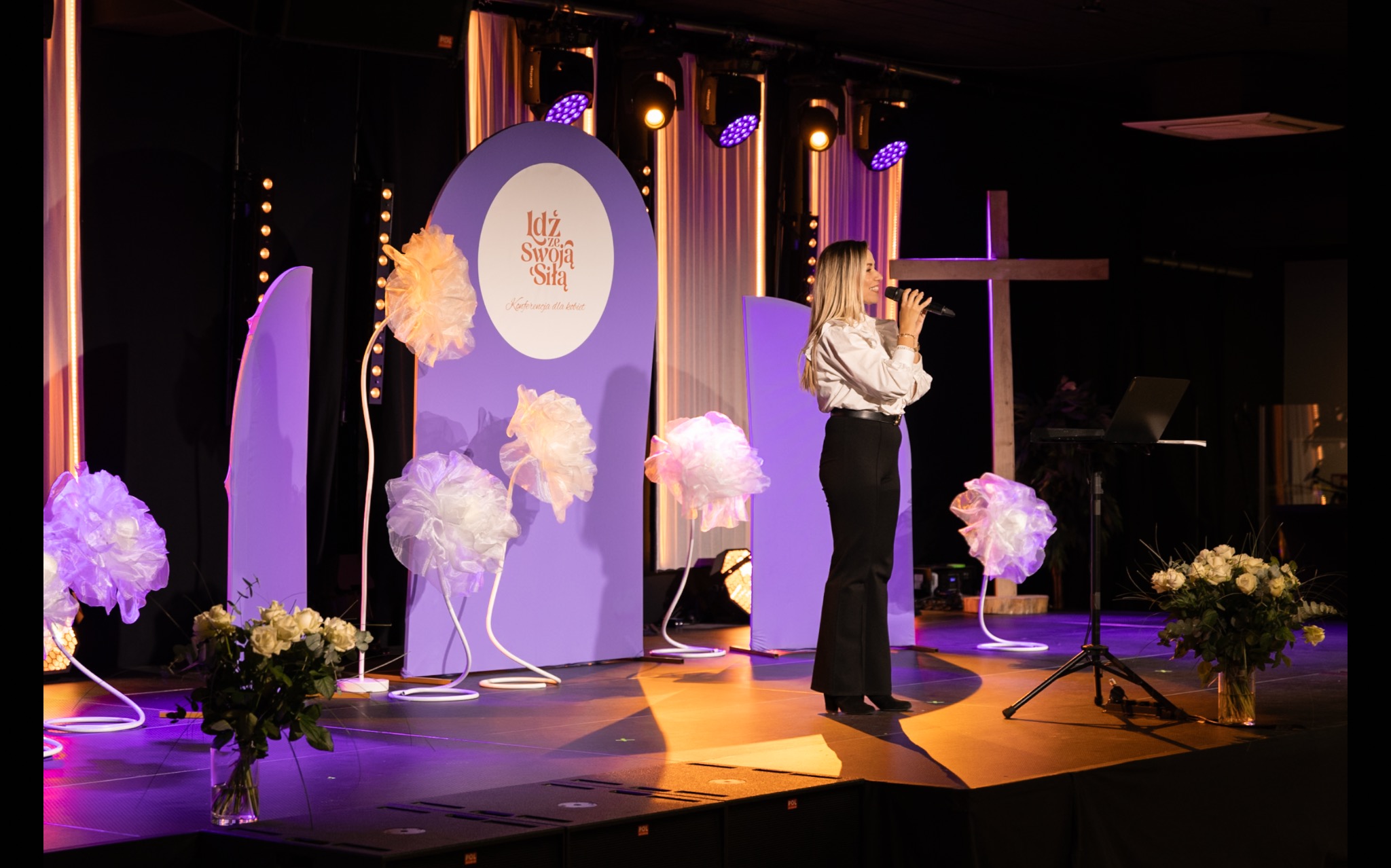 Kobieta śpiewa na scenie z dekoracjami w odcieniach fioletu i bieli, krzyż w tle