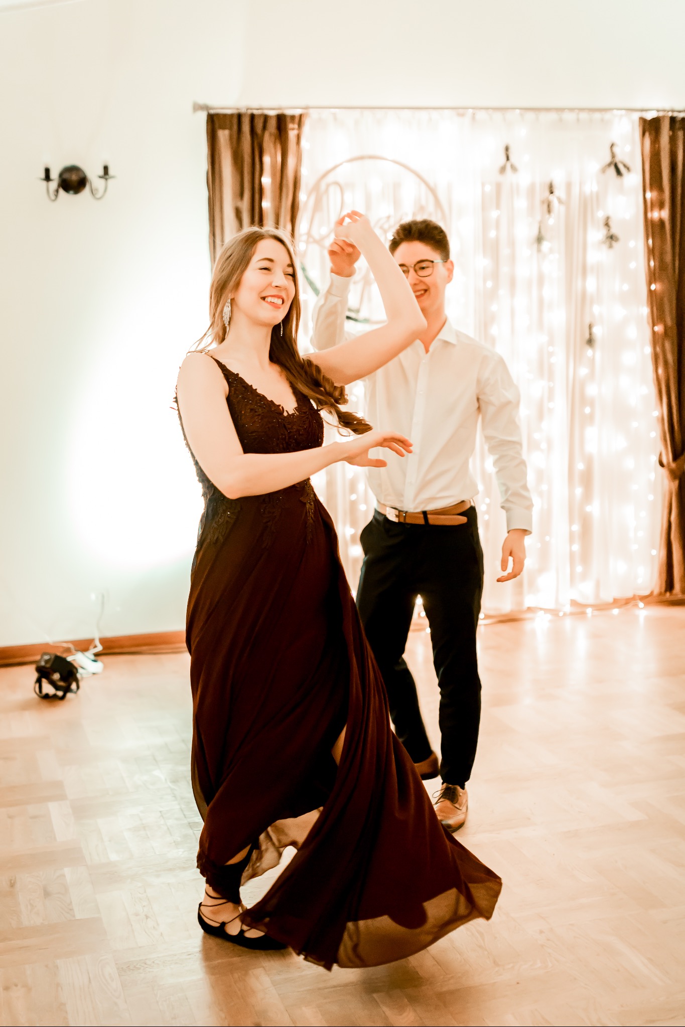 Uśmiechnięta para tańczy na parkiecie, kobieta w bordowej sukni z koronkową górą i baletkach, mężczyzna w białej koszuli i czarnych spodniach, w tle dekoracja ze światełek.